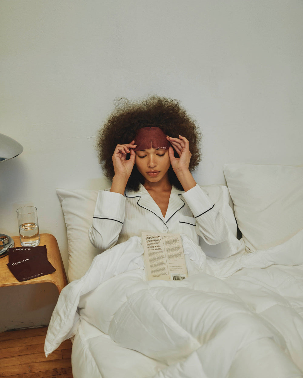 Woman sitting in bed placing a Revitalist15 self-heating eye mask over her eyes, demonstrating how to position and secure the ear loops.